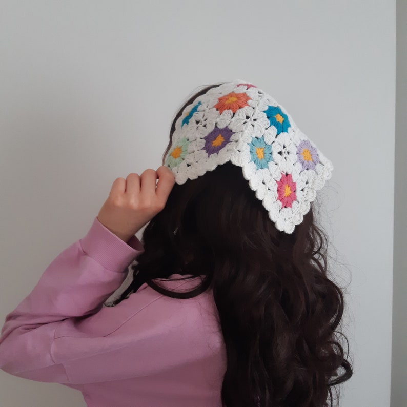 May include: A white crocheted headband with colorful flower accents. The headband is worn by a person with long, dark brown hair.