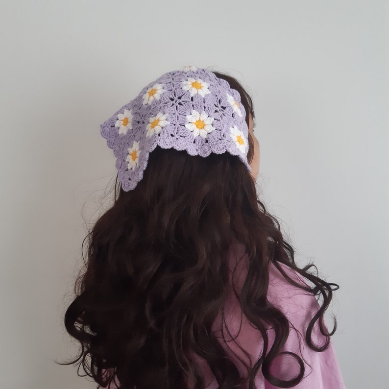 May include: A crocheted lavender bandana featuring a daisy pattern with white petals and yellow centers. The bandana is worn on the head, with the back of the head and long, dark brown hair visible.