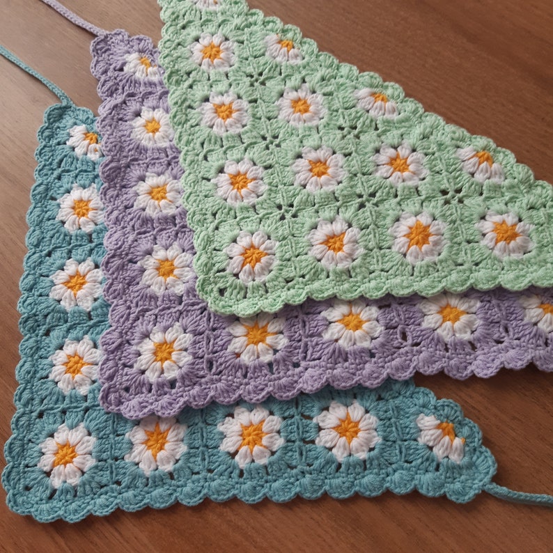 May include: Three crocheted triangle shawls in shades of green, purple, and blue with a daisy flower pattern.