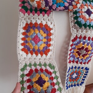 May include: A colorful crocheted scarf with a granny square pattern in shades of red, orange, yellow, green, blue, and purple. The scarf is made of a soft, textured yarn and has a fringe at the ends.