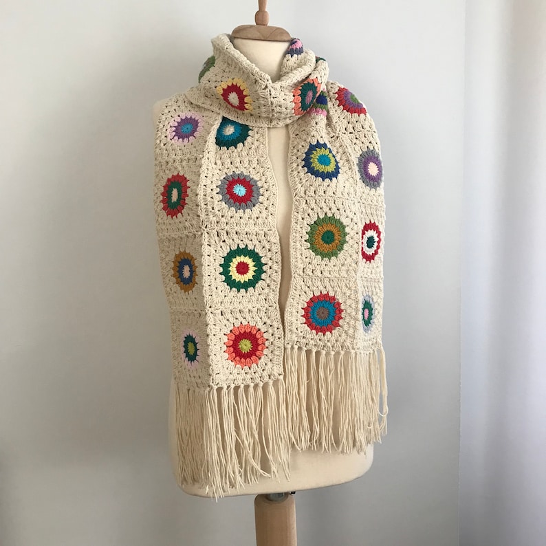 May include: A crocheted scarf with a fringe. The scarf is made of cream-colored yarn and features colorful circular motifs.