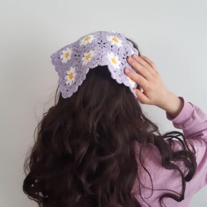 May include: A purple crocheted headband with white daisy flowers. The headband is worn on a person with long, dark brown hair.