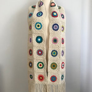 May include: A crocheted scarf with a fringe. The scarf is made of a cream-colored yarn and features colorful circles throughout.