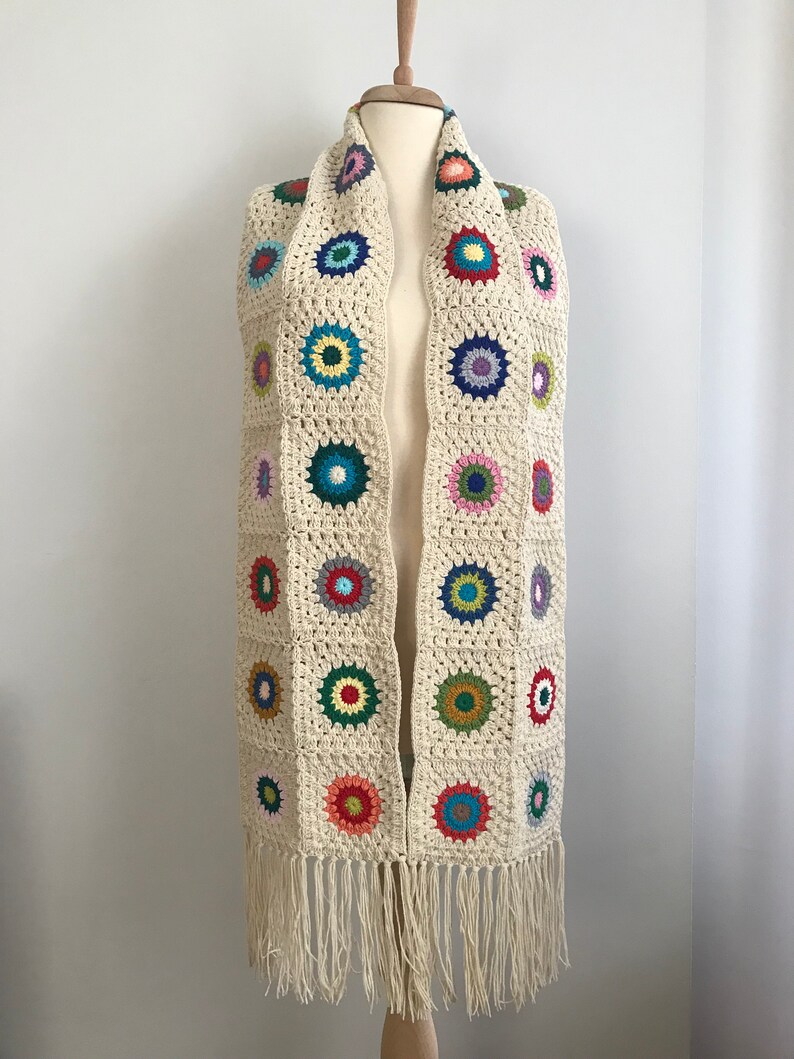 May include: A crocheted scarf with a fringe. The scarf is made of a cream-colored yarn and features colorful circles throughout.