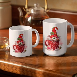 May include: Two white ceramic mugs with a Christmas gnome design. The gnome is wearing a red hat with white trim and is holding a candy cane. The mug on the left has a red and white snowflake design. The mug on the right has a red and white snowflake design with a red and white striped candy cane.
