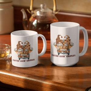 May include: Two white coffee mugs with a graphic of gnomes driving a truck with coffee cups and coffee beans. The text on the mugs reads "coffee lovin' chomies".