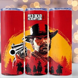 Puede incluir: Vaso rojo con un dise&ntilde;o gr&aacute;fico del videojuego Red Dead Redemption II. El dise&ntilde;o incluye un hombre con sombrero sosteniendo un arma, sobre un fondo de puesta de sol. El vaso es rojo con tapa plateada.