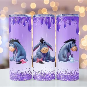 Puede incluir: Tres vasos t&eacute;rmicos morados con Eeyore de Winnie the Pooh. Cada vaso tiene un dise&ntilde;o de pintura goteando morada, un dibujo animado de Eeyore y una base brillante. El fondo est&aacute; borroso con luces bokeh.