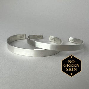 Puede incluir: Dos pulseras de puño plateadas de metal cepillado. Las pulseras tienen el texto "tailored metal" y "Sterling Silver 925" grabado en el interior. Un hexágono negro y dorado con el texto "NO GREEN SKIN" está en la esquina inferior derecha.
