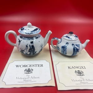 Peut inclure: Deux petites théières en porcelaine blanche avec des motifs bleus. L'une représente des personnages et des oiseaux, l'autre des personnages et un motif floral. Les deux sont présentées sur des cartes du Victoria & Albert Museum.