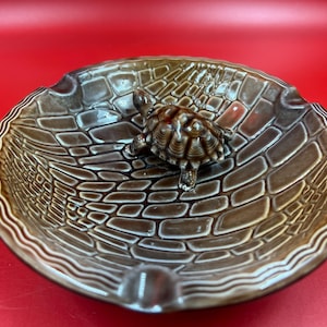 May include: A round brown ceramic ashtray featuring a raised, textured brick-like pattern on the interior surface. A small, three-dimensional brown turtle figurine is attached to the centre of the bowl. The rim has a wavy, grooved border.