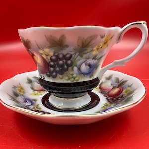May include: A vintage teacup and saucer set. The white porcelain cup and saucer feature a colorful fruit and floral design with gold trim. The cup has a black base and a white handle.