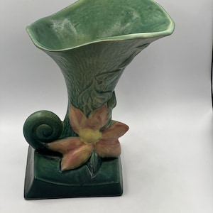 May include: A green glazed ceramic vase with a floral design. The vase is shaped like a trumpet with a curled base and a flower on the side.