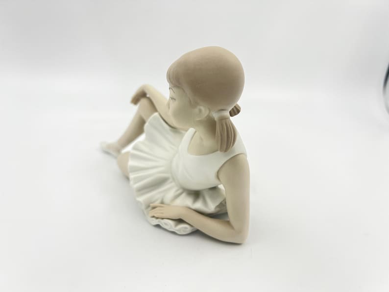 Puede incluir: Una figurita de porcelana de una ni&ntilde;a joven con un tut&uacute; blanco y sentada con las piernas cruzadas. Su cabello est&aacute; recogido en una coleta.