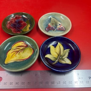 May include: Four small round ceramic dishes with raised botanical designs, including a red flower, a green leaf, a yellow leaf, and a multi-coloured leaf. A metal ruler is placed below the dishes, showing each dish is approximately 7.6 cm wide.