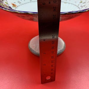 May include: A white porcelain pedestal bowl with a scalloped blue and gold rim, featuring a floral pattern. A metal ruler is positioned vertically against the bowl, showing a height measurement of approximately 10 cm from base to rim.
