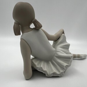 Puede incluir: Una figurita de porcelana de una joven bailarina que lleva un tut&uacute; blanco y est&aacute; sentada con las piernas cruzadas. Su cabello est&aacute; recogido en dos coletas.