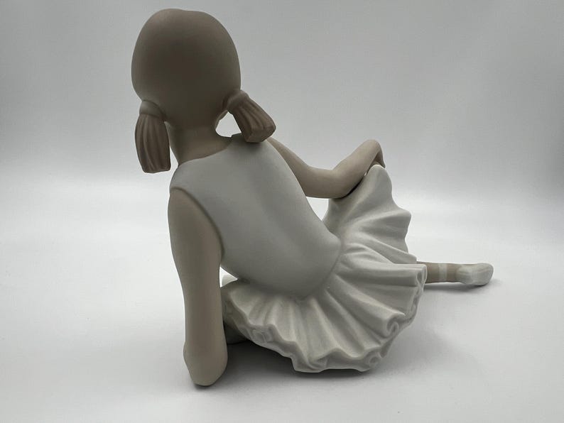 Puede incluir: Una figurita de porcelana de una joven bailarina que lleva un tut&uacute; blanco y est&aacute; sentada con las piernas cruzadas. Su cabello est&aacute; recogido en dos coletas.