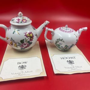 May include: Two white porcelain teapots with floral patterns, one larger with a lid and a curved handle, the other smaller. Both are displayed on cards from the Victoria & Albert Museum. The larger teapot has the word "BOW" on the card.