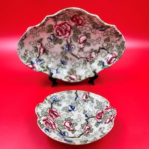 May include: Two vintage floral patterned ceramic serving dishes. The larger dish is oval shaped with a scalloped edge. The smaller dish is round with a scalloped edge. Both dishes have a white background with pink, green, and blue floral designs.