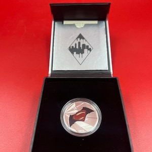 Peut inclure: Une pièce de monnaie argentée avec les logos de Superman et Batman, présentée dans un coffret noir. La pièce porte l'inscription "10 DOLLARS" et l'année "2016". Le coffret a un intérieur argenté et un motif de paysage urbain.