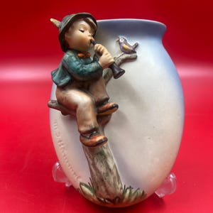 May include: A ceramic vase featuring a boy playing a flute, perched on a tree branch. The vase has a light blue and white gradient, with a small bird perched nearby. The boy wears a green jacket, brown pants, and a hat.