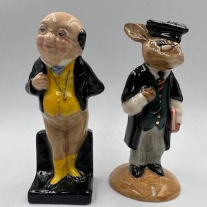 May include: A pair of bookends featuring a man and a rabbit. The man is wearing a yellow vest, brown pants, and a black jacket. The rabbit is wearing a black robe and a cap. Both bookends are standing on black bases.