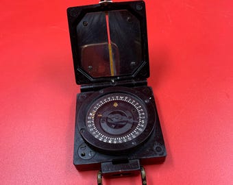 May include: A black compass with a hinged lid, open to show a circular dial with directional markings. The compass has a metal loop for carrying and a red and yellow sighting line. The background is a solid red.