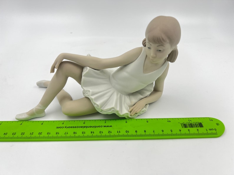 Puede incluir: Una figurita de porcelana de una ni&ntilde;a peque&ntilde;a con un tut&uacute; blanco y sentada con las piernas cruzadas. Tiene el pelo largo y casta&ntilde;o y mira hacia abajo.