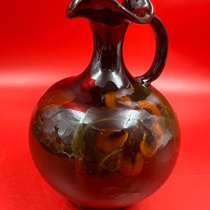 May include: A dark brown glass pitcher with a rounded body and a ruffled rim. The surface features an embedded floral pattern with orange and green tones visible through the translucent glass. It has a single curved handle on the side.