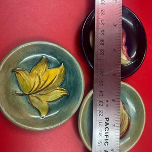 May include: Three circular ceramic dishes featuring embossed leaf designs. One dish is teal with a yellow leaf, while two are smaller with dark blue and light green glazes. A metal ruler shows the dishes measure approximately 9 cm in diameter.