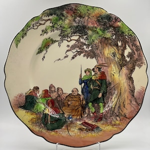 May include: A vintage ceramic plate with a hand-painted scene of a group of people gathered under a large tree. The people are dressed in medieval clothing and are listening to a man in a brown robe. The plate has a scalloped edge and is decorated with a black rim.