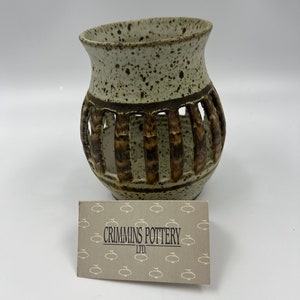 Peut inclure: Un vase en céramique mouchetée avec un design unique. Le vase présente des sections découpées verticales et un dessus évasé. La palette de couleurs comprend des nuances de marron et de beige. Le texte "CRIMMINS POTTERY LTD." est visible sur une carte devant le vase.
