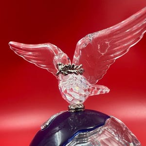 May include: A clear crystal dove figurine with outstretched wings, holding a silver olive branch in its beak. The dove sits atop a blue and clear crystal globe, set against a red background. The figurine is a decorative item.