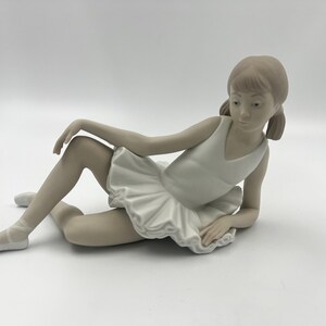 Puede incluir: Una figurita de porcelana de una joven bailarina en un tut&uacute; blanco, tumbada de lado con una pierna extendida y la otra doblada. Tiene una expresi&oacute;n facial delicada y el pelo recogido en una coleta.