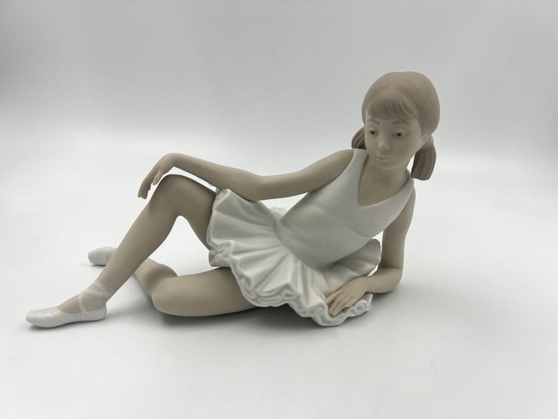 Puede incluir: Una figurita de porcelana de una joven bailarina en un tut&uacute; blanco, tumbada de lado con una pierna extendida y la otra doblada. Tiene una expresi&oacute;n facial delicada y el pelo recogido en una coleta.