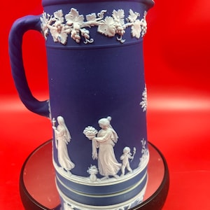 May include: A matte cobalt blue ceramic jug featuring white relief ornamentation. The upper rim displays a grape vine motif, while the main body depicts classical figures and a small child in white relief against the solid blue background.