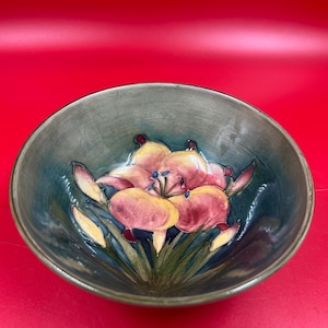 May include: A round ceramic bowl featuring a hand-painted floral design at the centre. The interior displays a large, multi-petaled flower in shades of pink and yellow with green stems, set against a deep blue and grey glazed background.