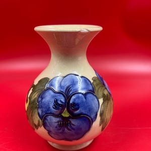 May include: A cream-coloured ceramic vase with a bulbous body and a flared rim. The surface features a hand-painted design of large blue pansy flowers with green leaves. There is a small chip visible on the upper edge of the vase rim.