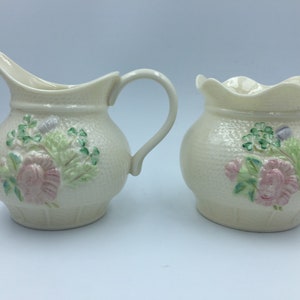 May include: A set of two cream-coloured ceramic creamer and sugar containers with floral and shamrock designs.