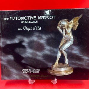 Peut inclure: Un livre intitulé "The Automotive Mascot Worldwide" avec une sculpture de couleur bronze sur la couverture. La sculpture représente une figure avec les bras levés. La couverture du livre est noire avec du texte blanc et les mots "un Objet d'Art".
