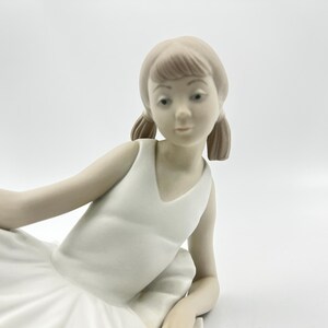 Puede incluir: Una figurita de porcelana de una ni&ntilde;a joven con un vestido blanco y un tut&uacute; de ballet. Tiene el pelo casta&ntilde;o y est&aacute; sentada con las piernas cruzadas.
