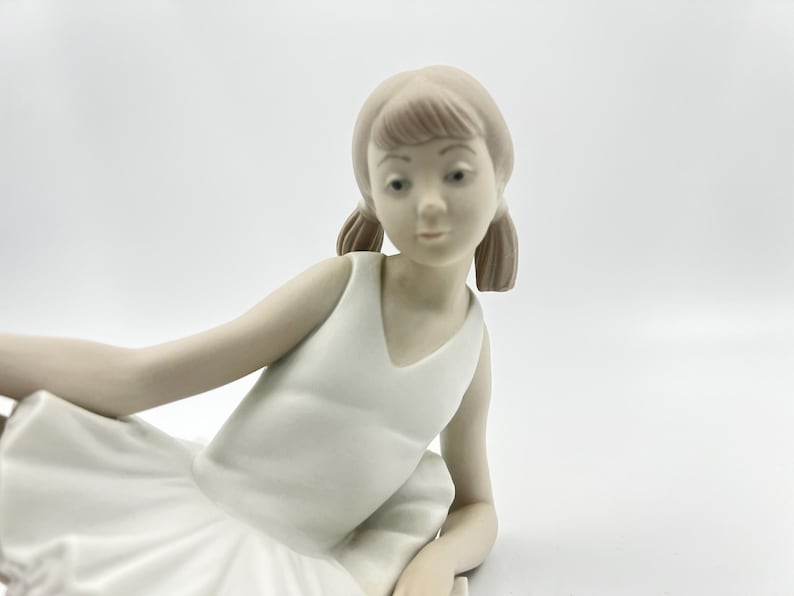Puede incluir: Una figurita de porcelana de una ni&ntilde;a joven con un vestido blanco y un tut&uacute; de ballet. Tiene el pelo casta&ntilde;o y est&aacute; sentada con las piernas cruzadas.