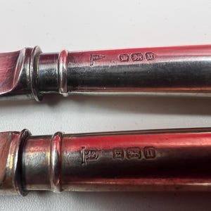 Peut inclure: Gros plan sur deux couteaux en argent antiques. Les manches sont de couleur argent brillant, avec une bande décorative près de la lame. Les lames présentent des signes d'usure. Chaque manche porte des marques estampillées.