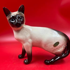 May include: A ceramic Siamese cat figurine with a cream-colored body and dark brown markings on the face, ears, paws, and tail. The cat has blue eyes and is posed in a seated position. A small green sticker is on the side.