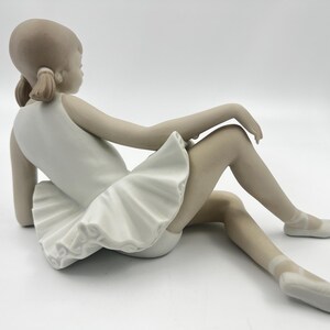 Puede incluir: Una figurita de porcelana de una joven bailarina que lleva un tut&uacute; blanco y zapatillas de punta. Est&aacute; sentada con las piernas extendidas y los brazos apoyados en las rodillas.