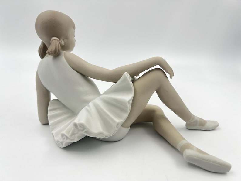 Puede incluir: Una figurita de porcelana de una joven bailarina que lleva un tut&uacute; blanco y zapatillas de punta. Est&aacute; sentada con las piernas extendidas y los brazos apoyados en las rodillas.