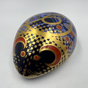 May include: A ceramic egg-shaped ornament with a blue, black, gold, and red geometric pattern. The ornament features a chequered pattern, crescent moons, and stylised eyes.