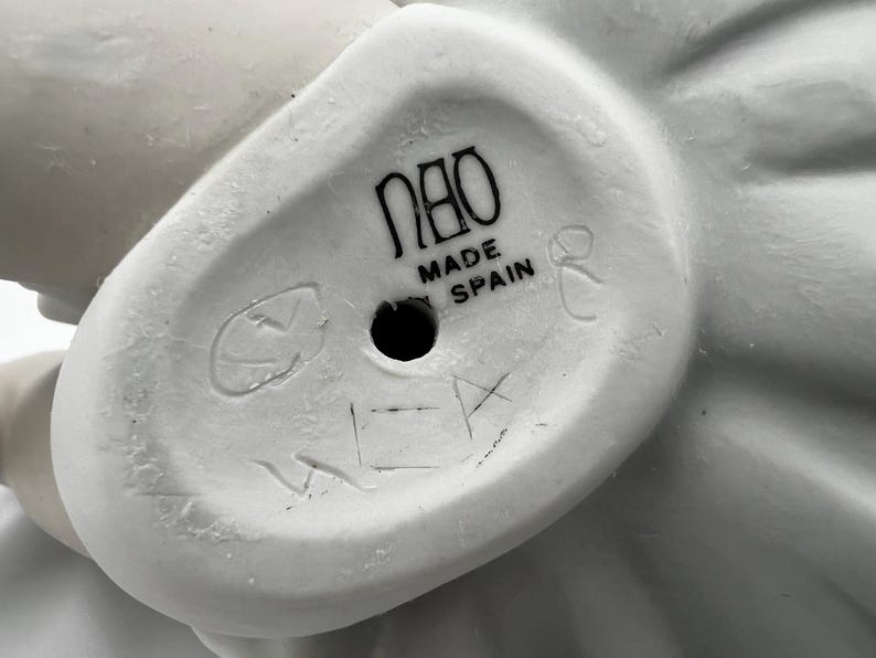 Puede incluir: Figurita de porcelana blanca con un dise&ntilde;o detallado. La base de la figurita tiene el texto "Nao Made in Spain" y un peque&ntilde;o agujero.