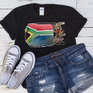 Puede incluir: Camiseta negra con un dise&ntilde;o de acuarela de la bandera sudafricana, con el texto "South Africa". La camiseta se combina con pantalones cortos de mezclilla y zapatillas de lona grises. El dise&ntilde;o incluye un tambor y otros instrumentos musicales.